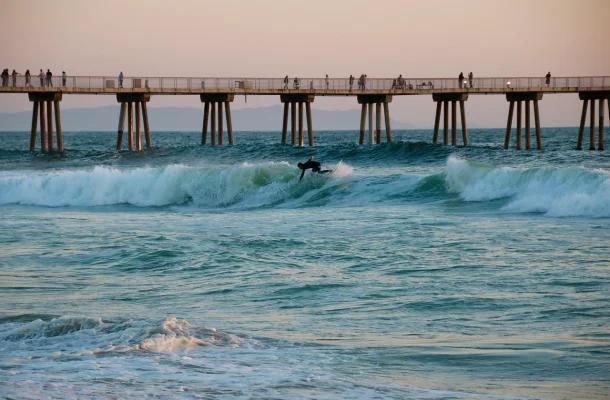 Ocean waves crash near a wooden pier with people fishing at sunset in Los Angeles coastal area
