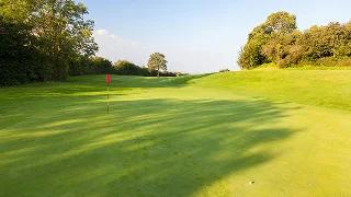 Pristine golf course green with flag and rolling fairways surrounded by mature trees under clear sky