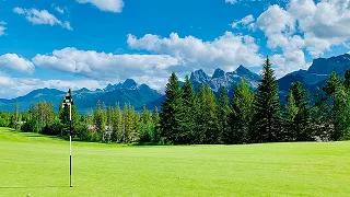 Pristine golf course with manicured green fairway and flag, surrounded by dense pine forests and dramatic mountain peaks under blue sky