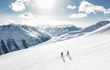 Two skiers descending pristine powder slopes with snow-capped mountain peaks under bright alpine sun