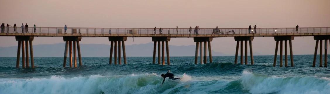 Surfer riding waves beneath wooden pier with spectators watching from above during golden hour